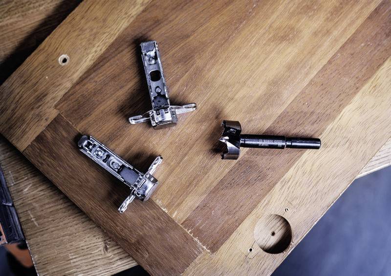 Detailný záber mechanických pántov a vŕtačky na drevenej doske, pravdepodobne v kontexte montáže nábytku.