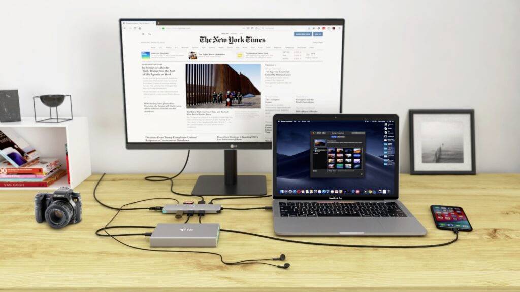 Písací stôl s laptopom pripojený k monitoru, na ktorom je zobrazený 'The New York Times'. Vedľa leží fotoaparát a smartfón.