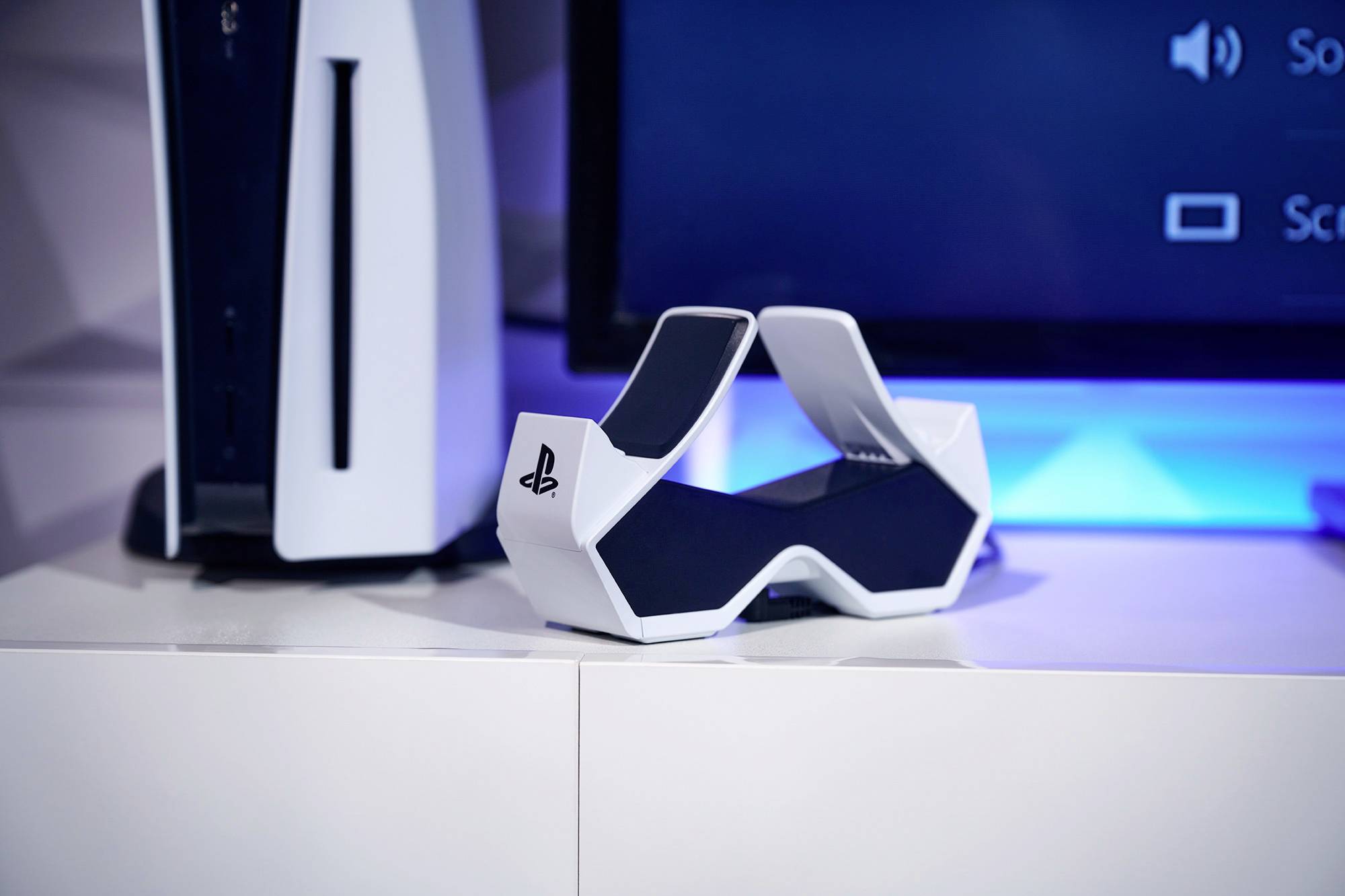 Herná konzola PlayStation 5 a biela nabíjacia stanica PS5 na polici pred televízorom.