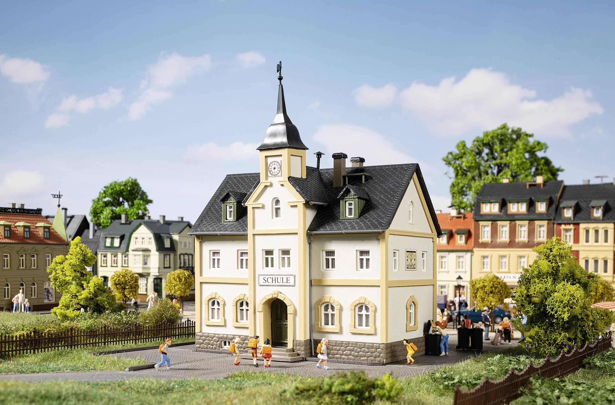 Miniatúrna dedina s modelom školy v centre. Ľudské figúrky oživujú scénu v útulnej dedinskej krajine.
