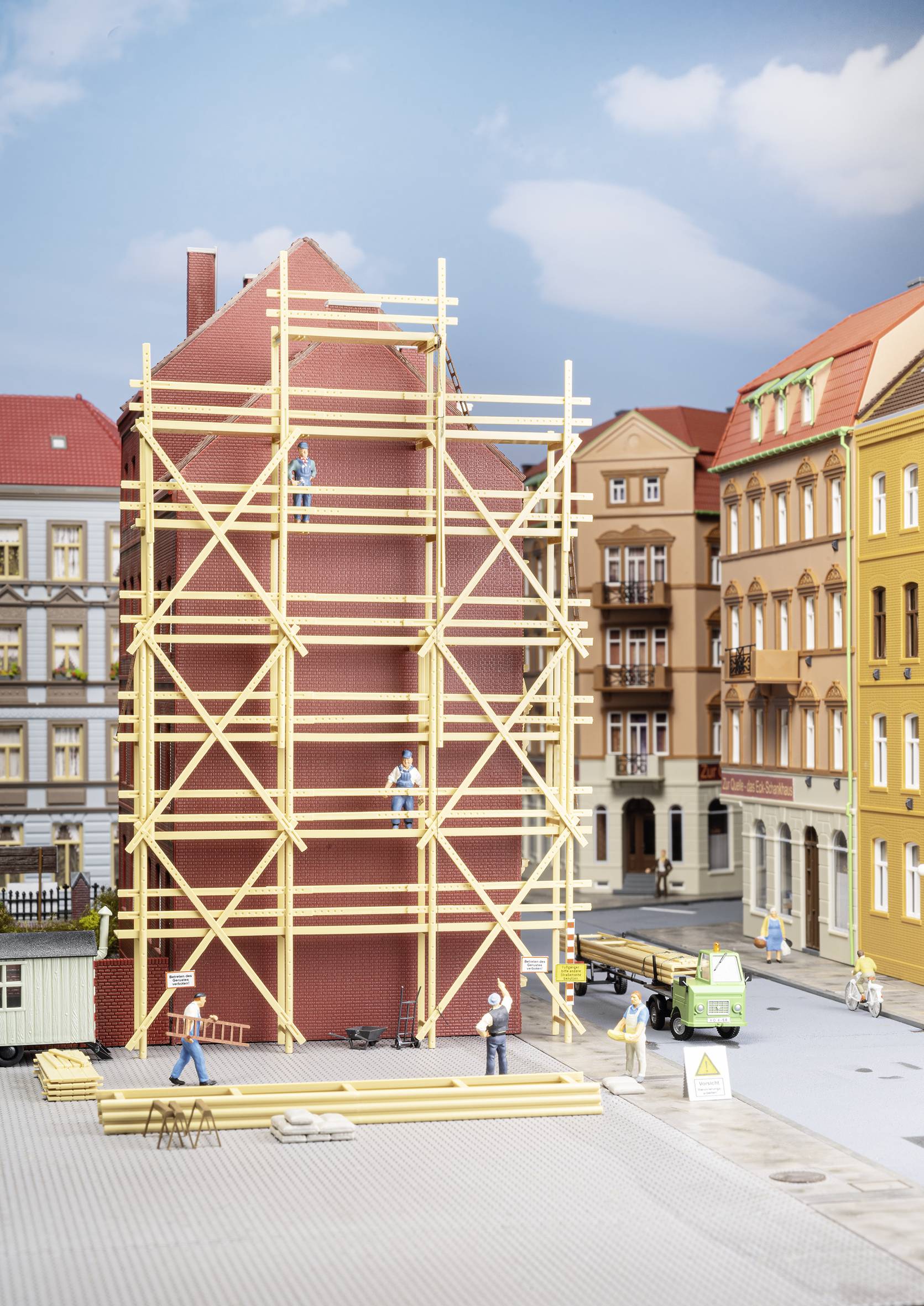 Model staveniska v meste s budovou na lešení, robotníkmi a malým nákladným autom v popredí.