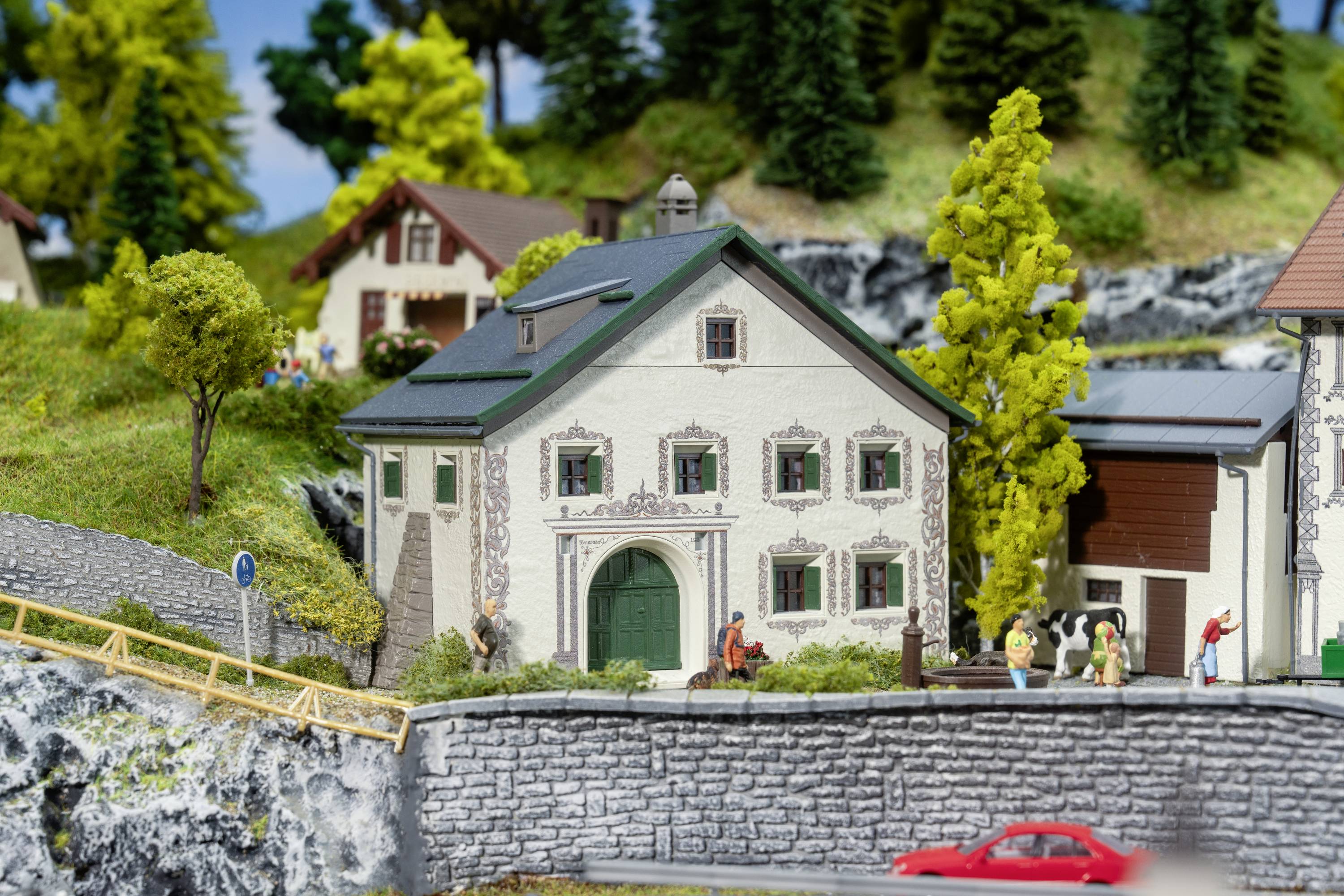 Miniatúrna dedina s hrázdeným domom, zelenými stromami a figúrkami. Ľudia sa prechádzajú po chodníku, dole prechádza červené auto.