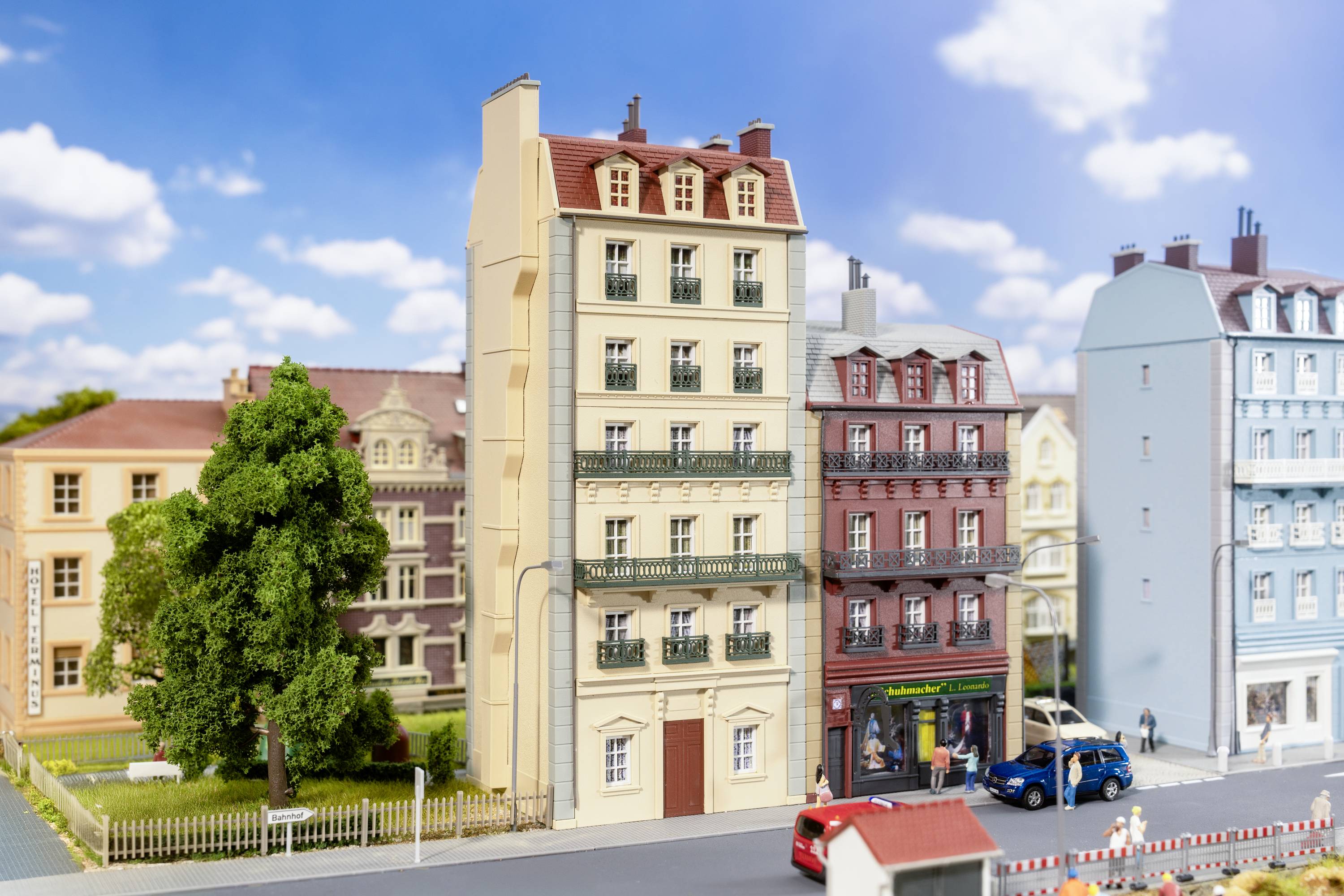 Miniaturná mestská scéna s viacerými detailne spracovanými budovami, autami a chodcami na ulici pod modrým nebom s oblakmi.