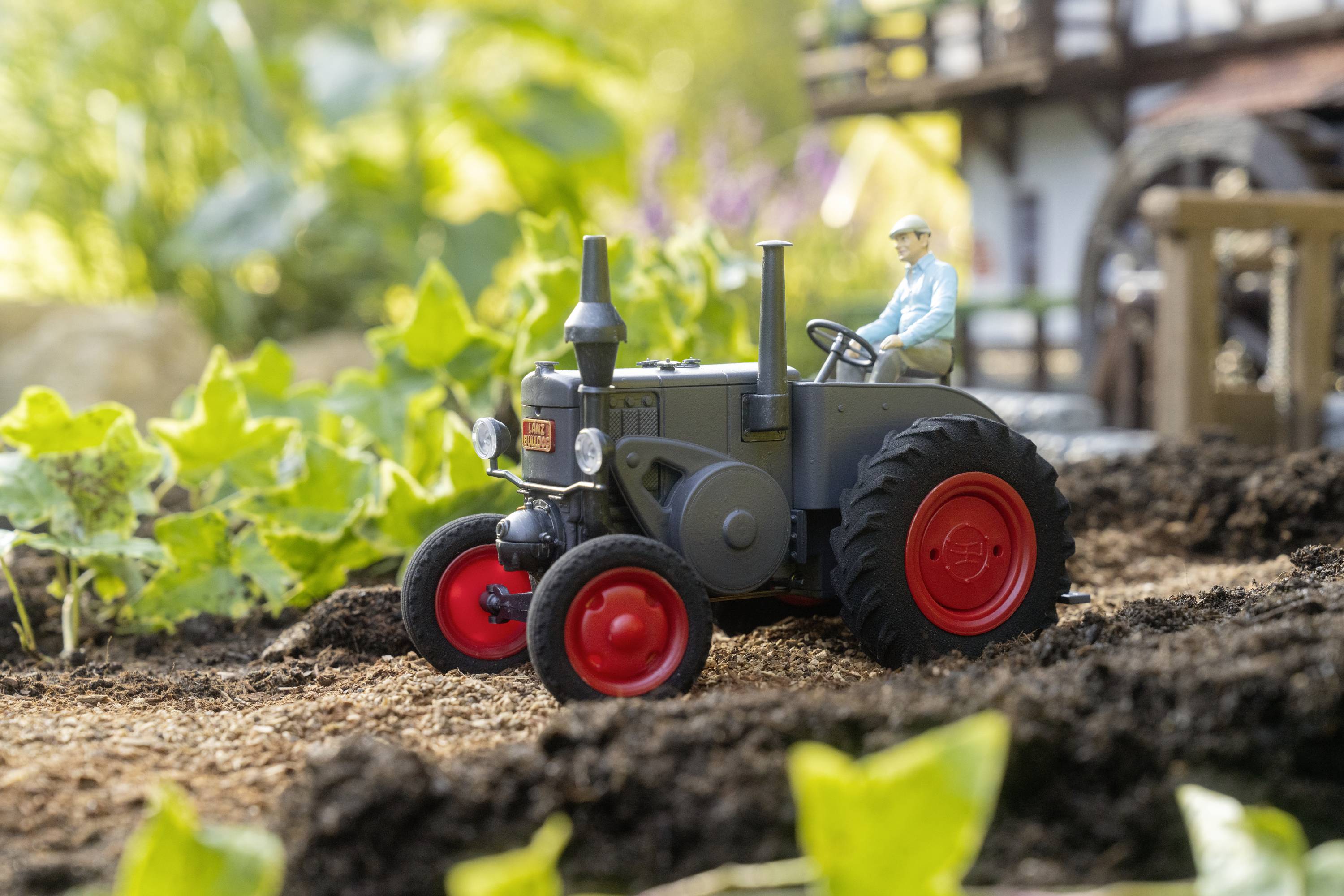 Malý model traktora s vodičom, ktorý jazdí cez miniatúrnu záhradu s rastlinami a zemou.