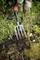 Osoba prekopáva pôdu záhradnými vidlami v záhrade. Okolo sú viditeľné zelené rastliny a gumené čižmy.