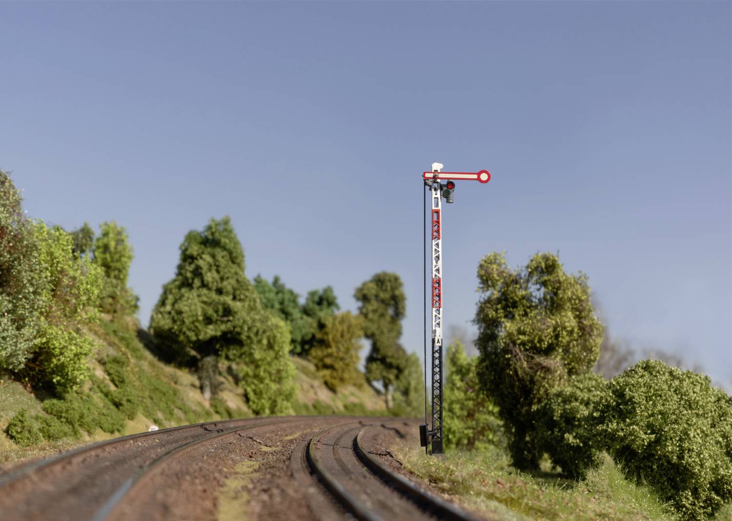Model elektrického vláčika s traťou zakrivujúcou sa cez krajinu, s mechanickým návestným stĺpom, obklopeným stromami a hustou vegetáciou.