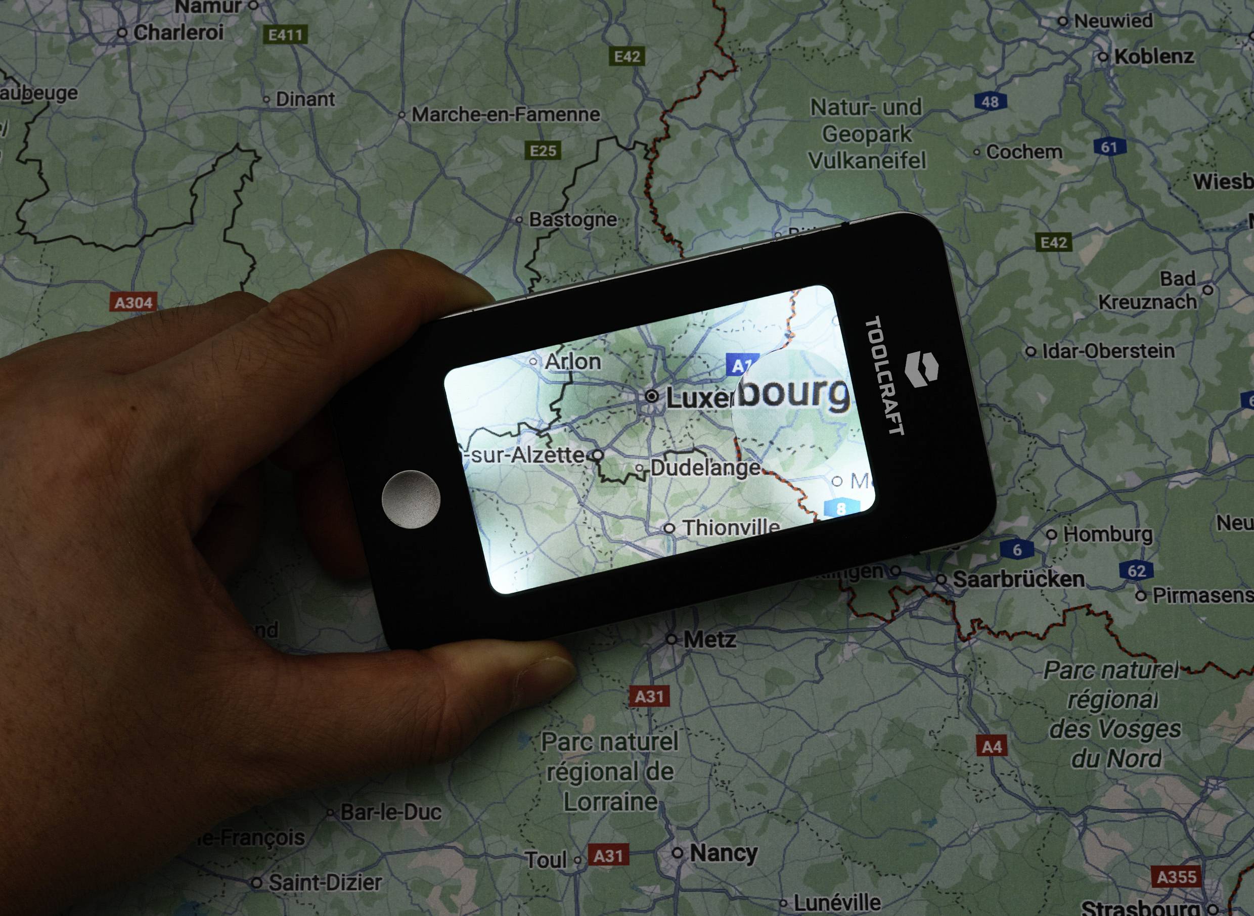 Ruka drží digitálnu lupu nad mapou zobrazujúcou región Luxemburska, pričom zvýrazňuje viditeľnosť názvov miest ako 'Luxembursko' a 'Arlon'.