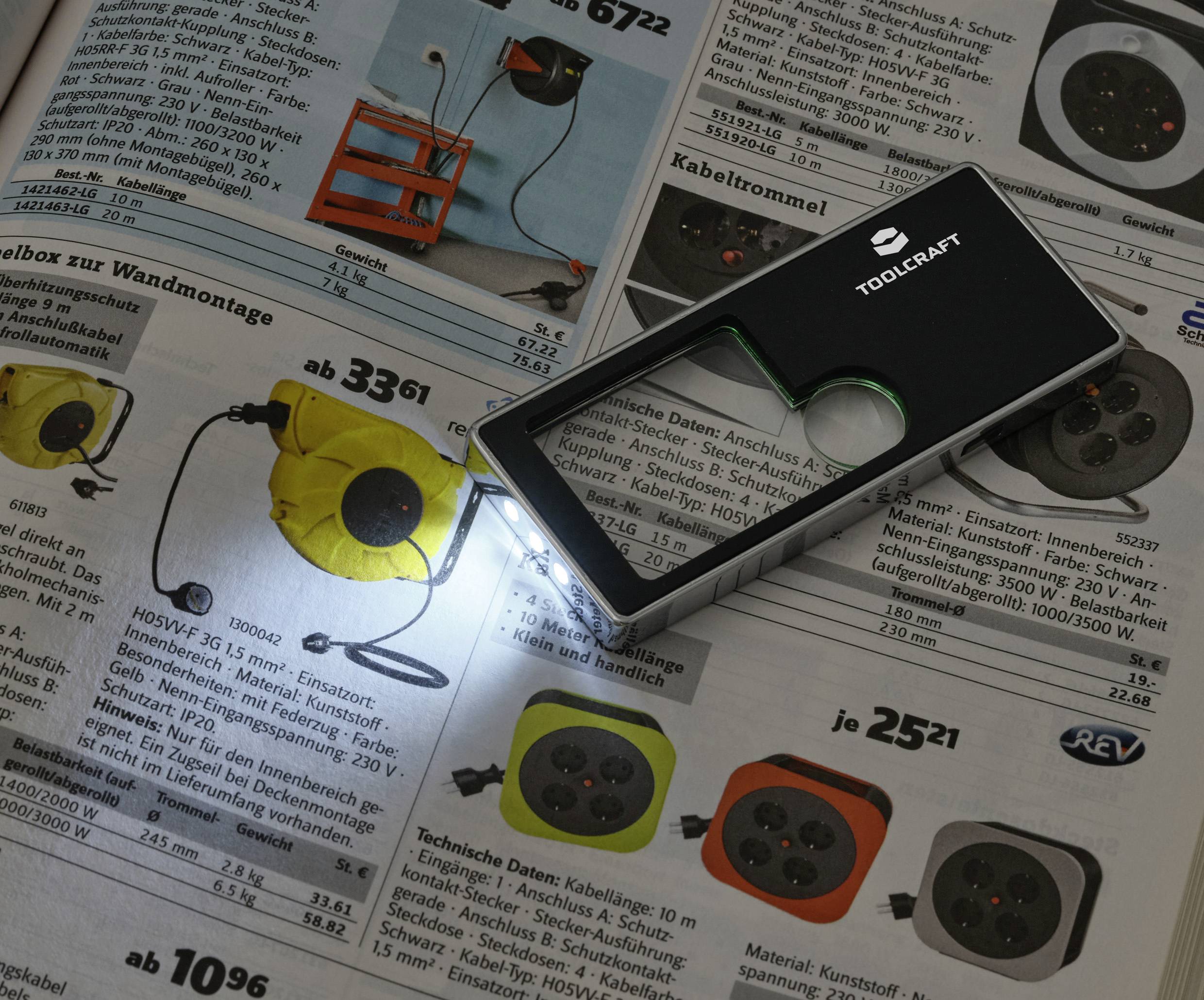 Obdĺžnikový čierny lup s LED svetlom, umiestnený nad katalógovou stránkou zobrazujúcou elektrické zásuvky a predlžovacie káble.