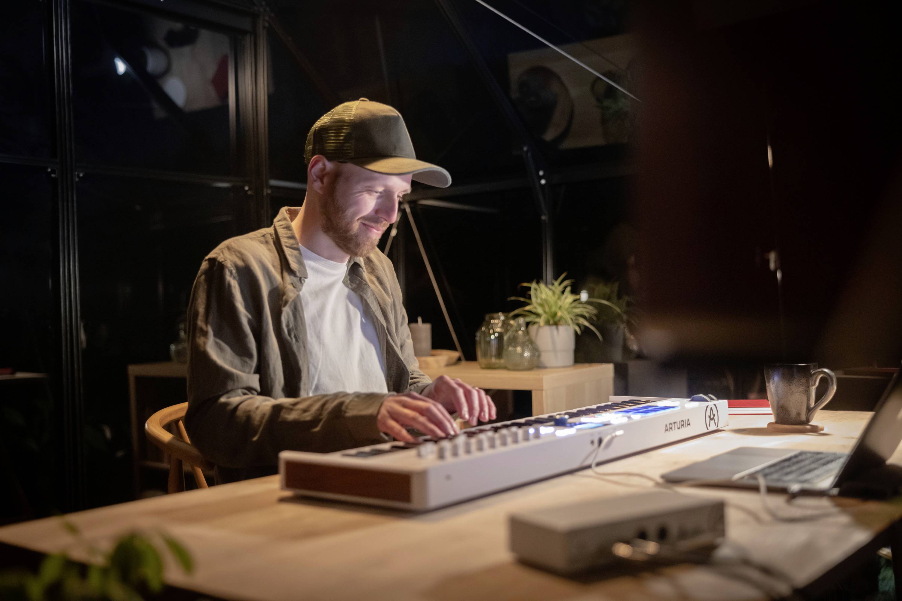 Muž s čiapkou hrá v zatemnenej miestnosti na elektronickom klaviatúre. Vľavo stojí šálka a rastlina.