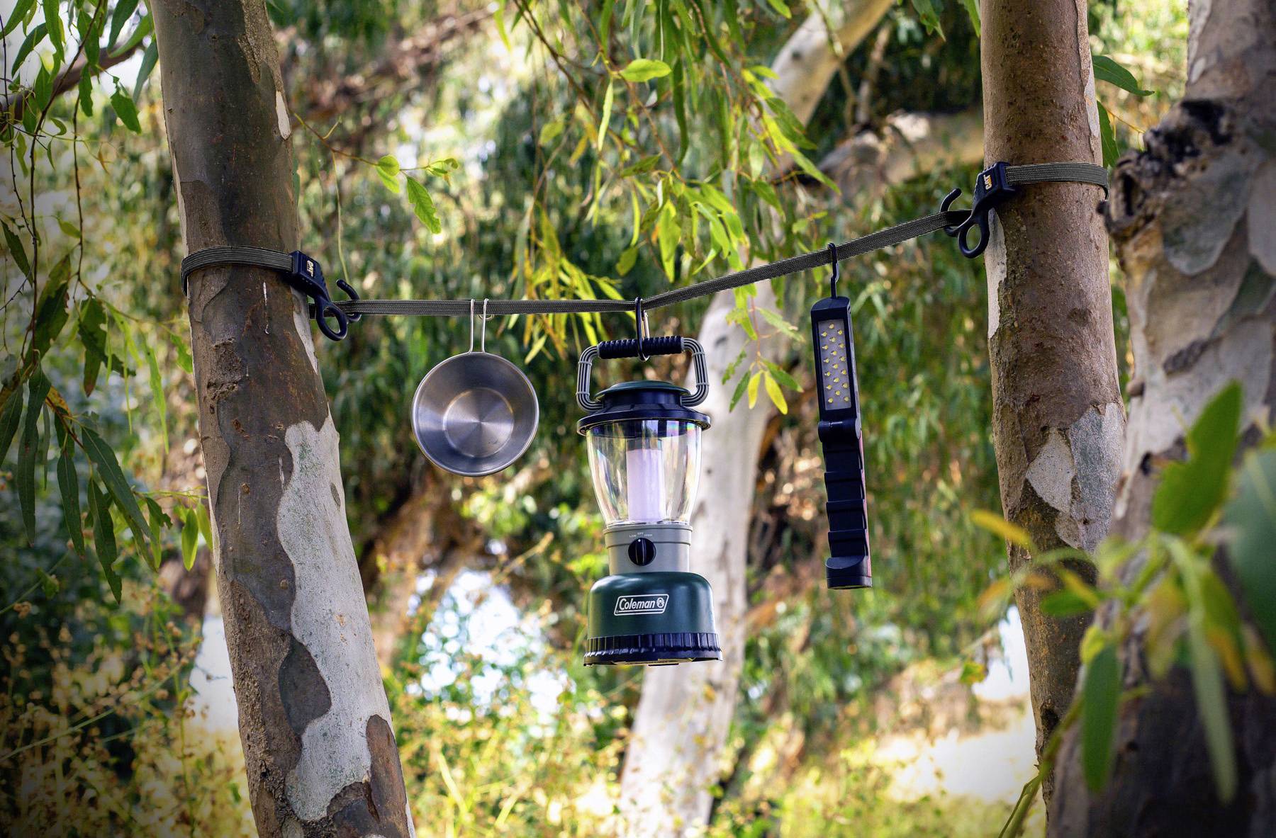 Kempingová lampa, hrniec a baterka visia na popruhu medzi dvoma stromami v lese. Slnečné svetlo presvitá cez listnaté konáre.