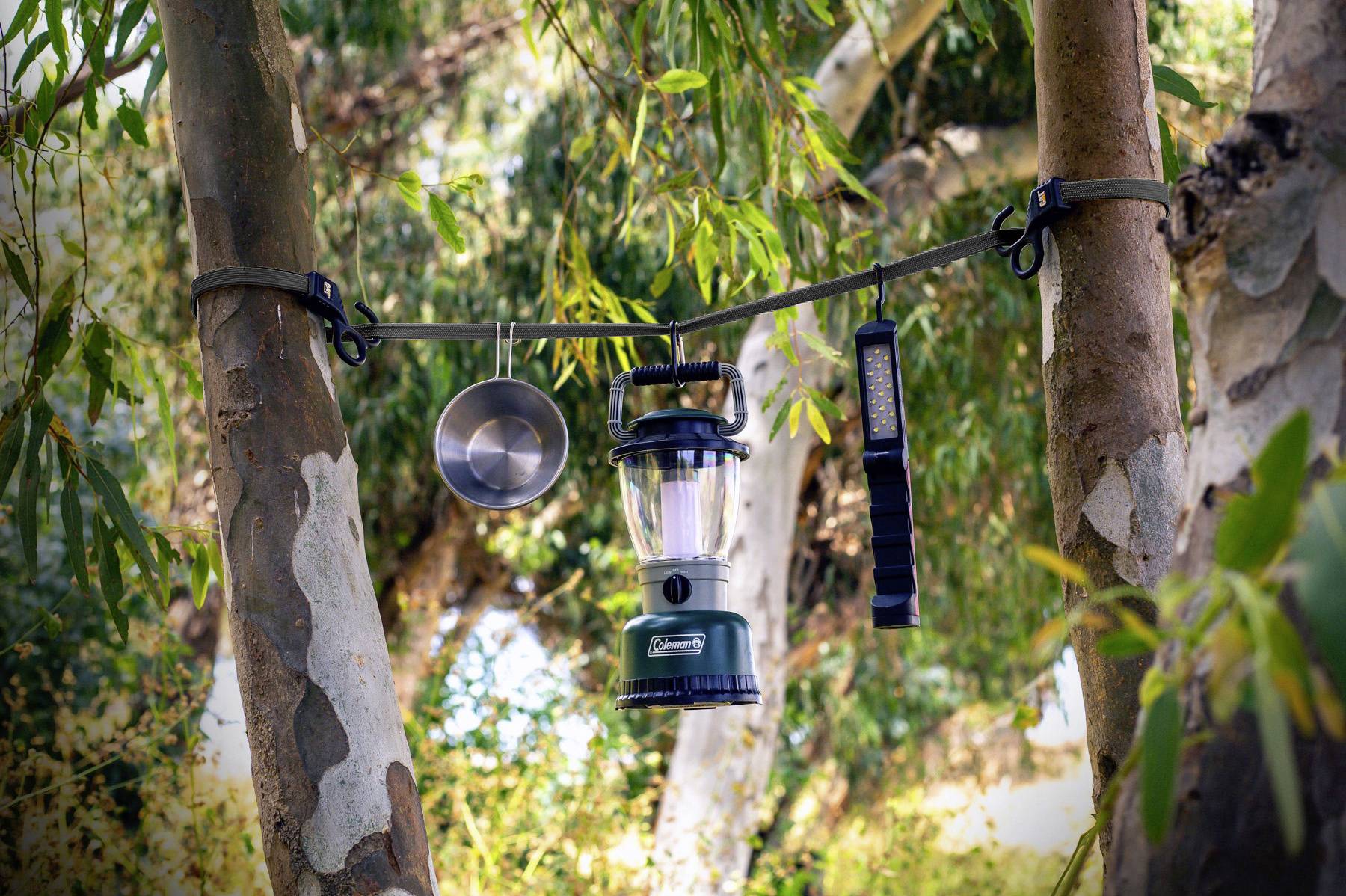 Kempingová lampa a iné nástroje visia na lane medzi dvoma stromami v lese, obklopené hustými zelenými listami.