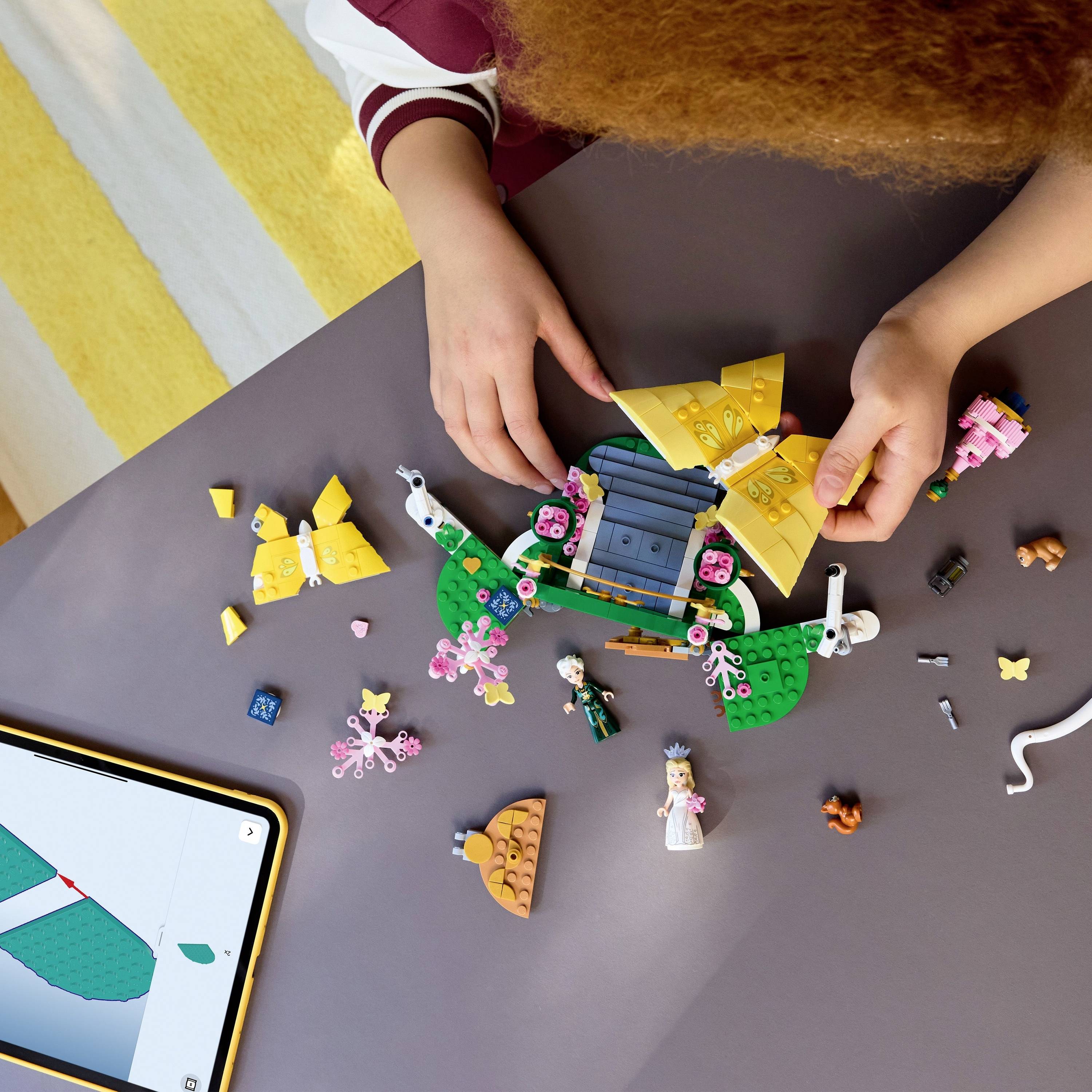 Dieťa zostavuje LEGO súpravu z rôznych farebných dielikov, vrátane figúrok a vozidiel, na stole vedľa digitálneho tabletu zobrazujúceho LEGO aplikáciu.