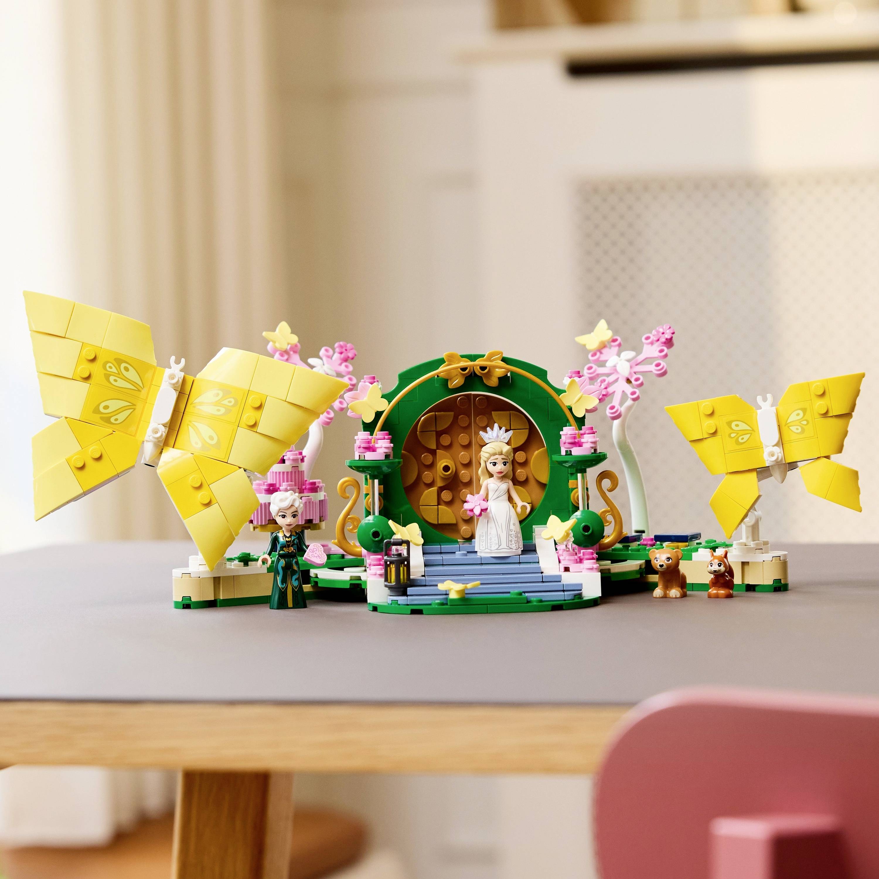 Scéna rozprávkovej krajiny z LEGO stavebnice s princeznou na modrej platforme, žltými motýľmi a malými figúrkami šaša a leva v blízkosti dekoratívnych rastlín.