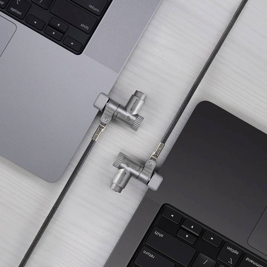 Dva laptopy sú zabezpečené káblovou zámkou na drevenom stole, čo zdôrazňuje dôraz na bezpečnosť a ochranu elektronických zariadení.