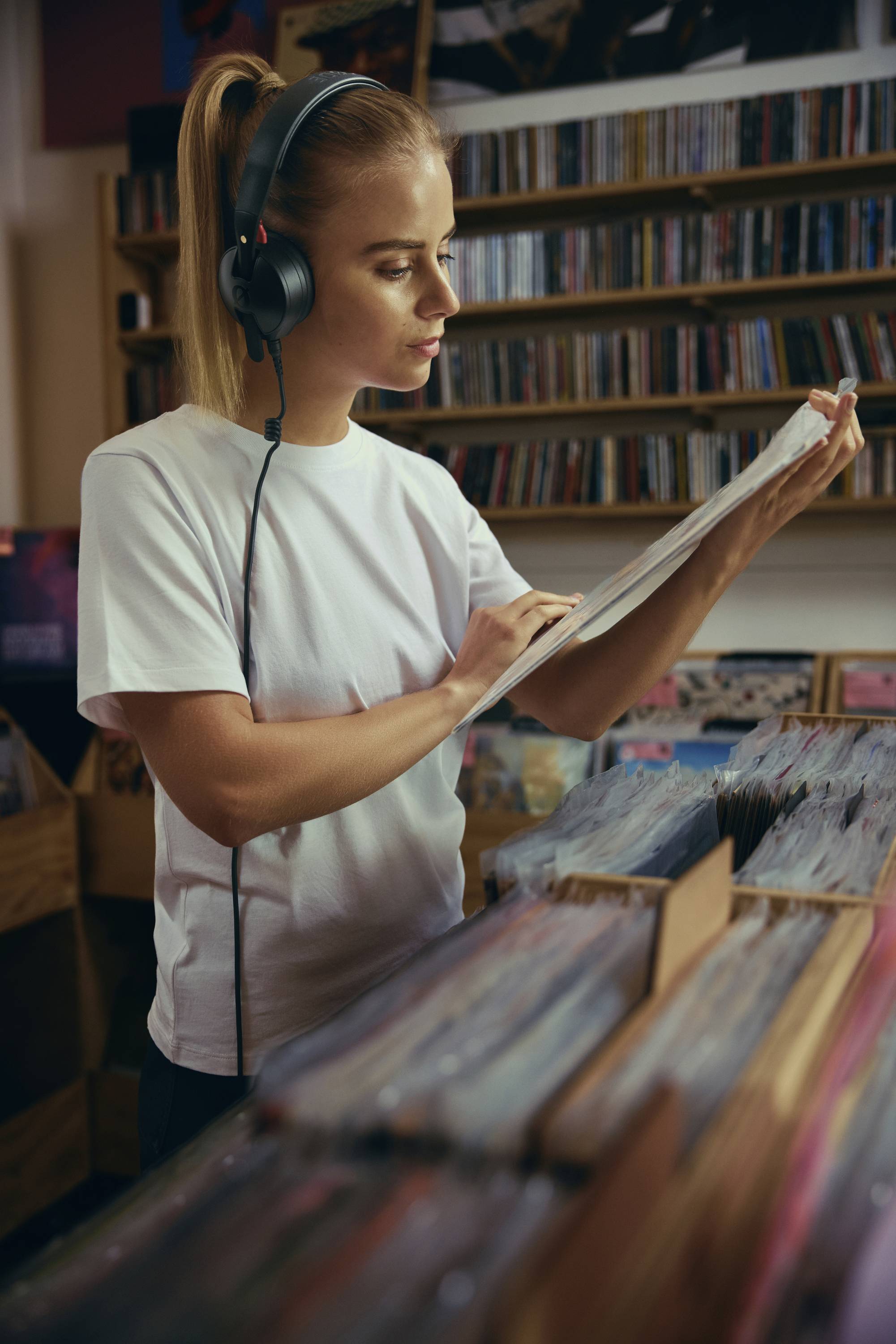 Osoba s nasadenými slúchadlami prezerá vinylové platne v hudobnom obchode, obklopená políčkami plnými albumov.