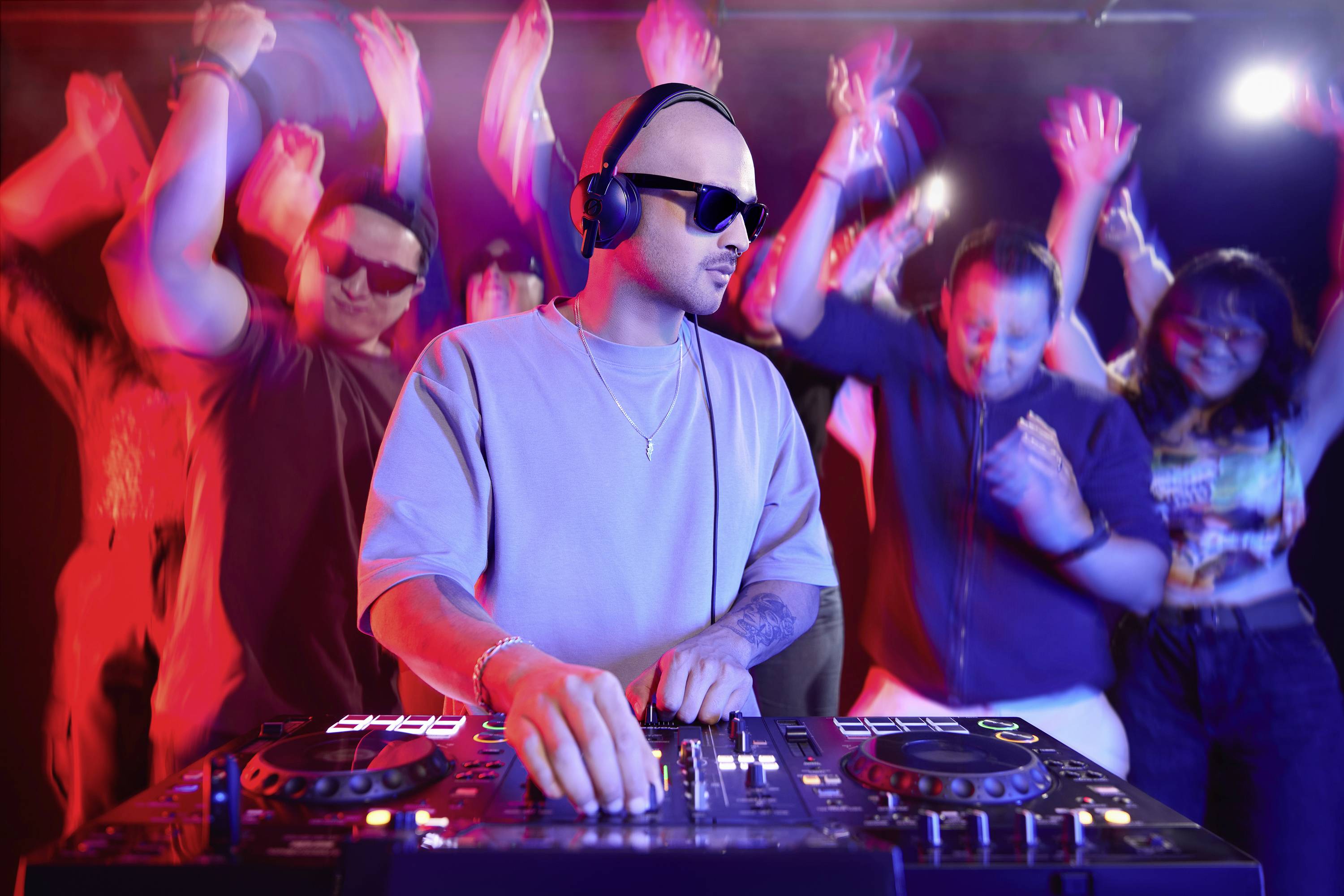 DJ so slúchadlami mixuje hudbu na konzole v živom klube. Ľudia s zdvihnutými rukami energicky tancujú v pestrofarebných svetlách.