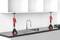 'Dva červené dverové zdviháky podopierajú bielu závesnú skrinku nad drezom a batériou, pričom na pracovnej doske je vedľa vŕtačka.'