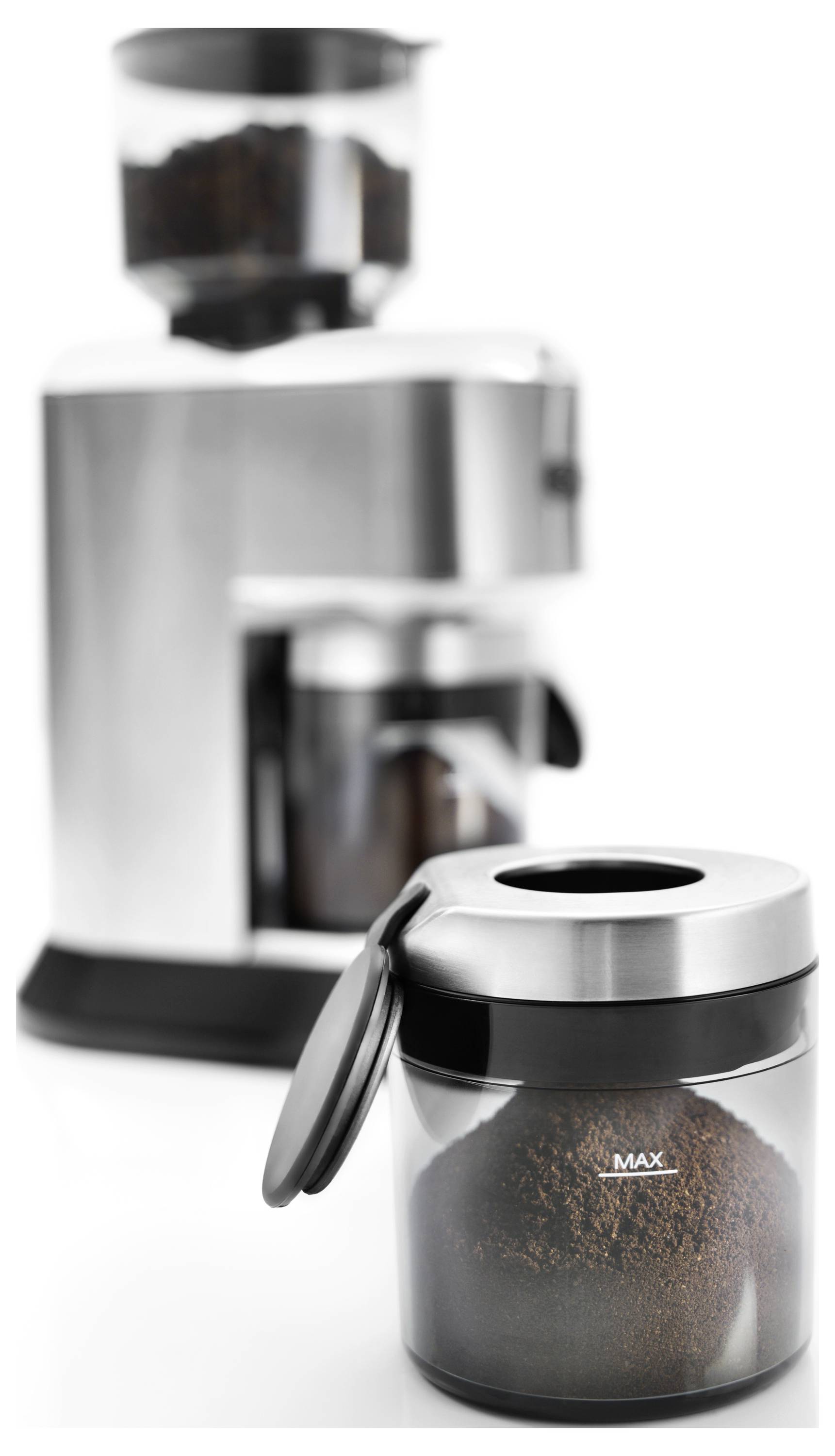 Priehľadná nádoba naplnená mletou kávou s označením "MAX" sa nachádza v popredí. V pozadí je mlynček na kávu s ďalšími kávovými zrnami.