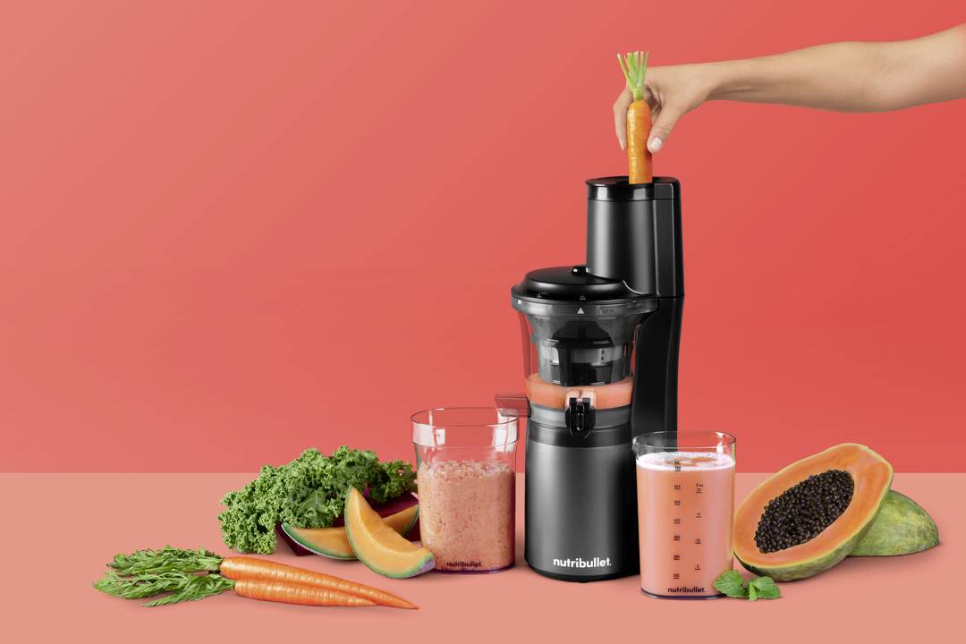 Ruka vkladá mrkvu do mixéra Nutribullet obklopená listami kelové kapusty, mrkvami, papájou a pohármi pomarančového džúsu na červenom pozadí.