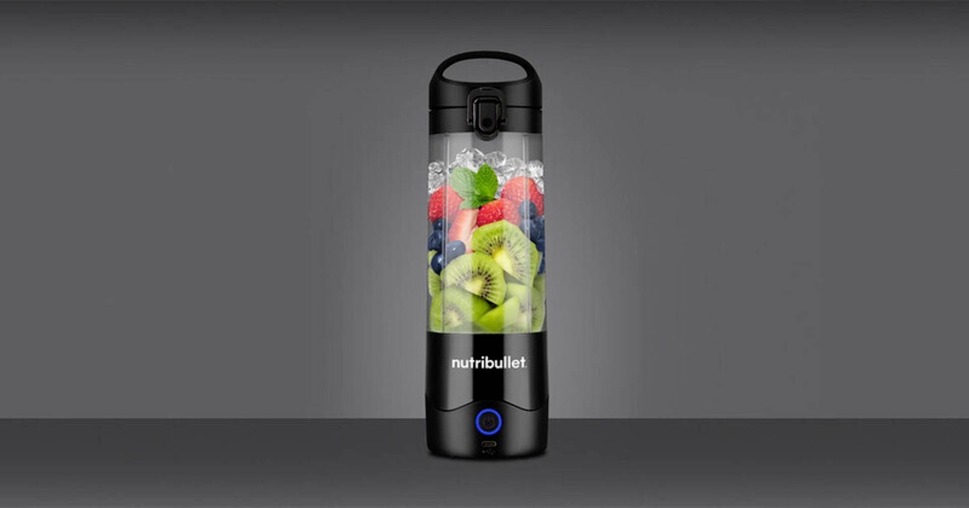 Prenosná fľaša mixéra naplnená zmiešanými ovocnými druhmi, vrátane kiwi, jahôd a čučoriedok, na šedivom pozadí.