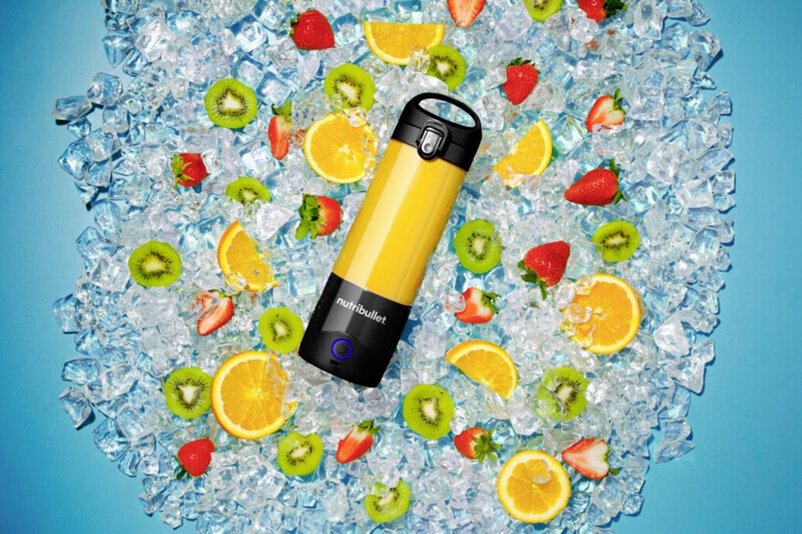 Jasne žltý mixér NutriBullet obklopený ľadom a nakrájaným ovocím vrátane pomarančov, kiwi, jahôd a limetiek na modrom pozadí.