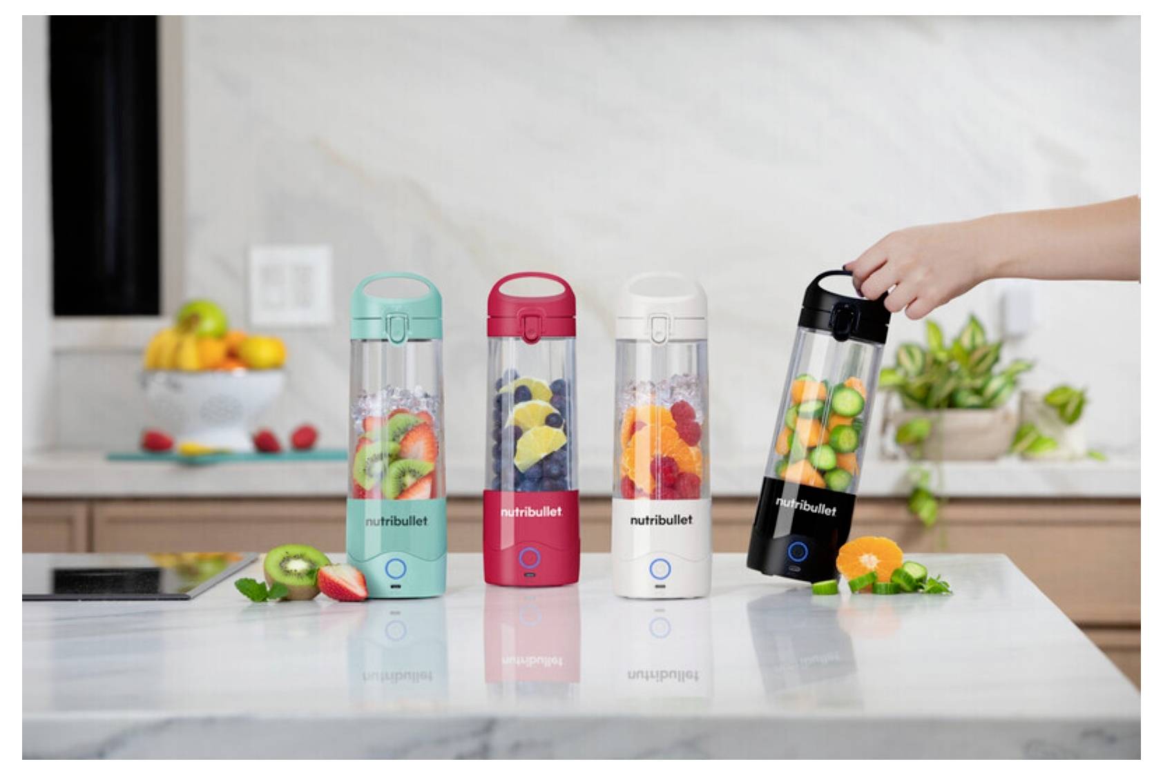Štyri farebné prenosné smoothie mixéry naplnené ovocím, stojace na kuchynskej linke. Ruka drží čierny mixér, pripravený na použitie.