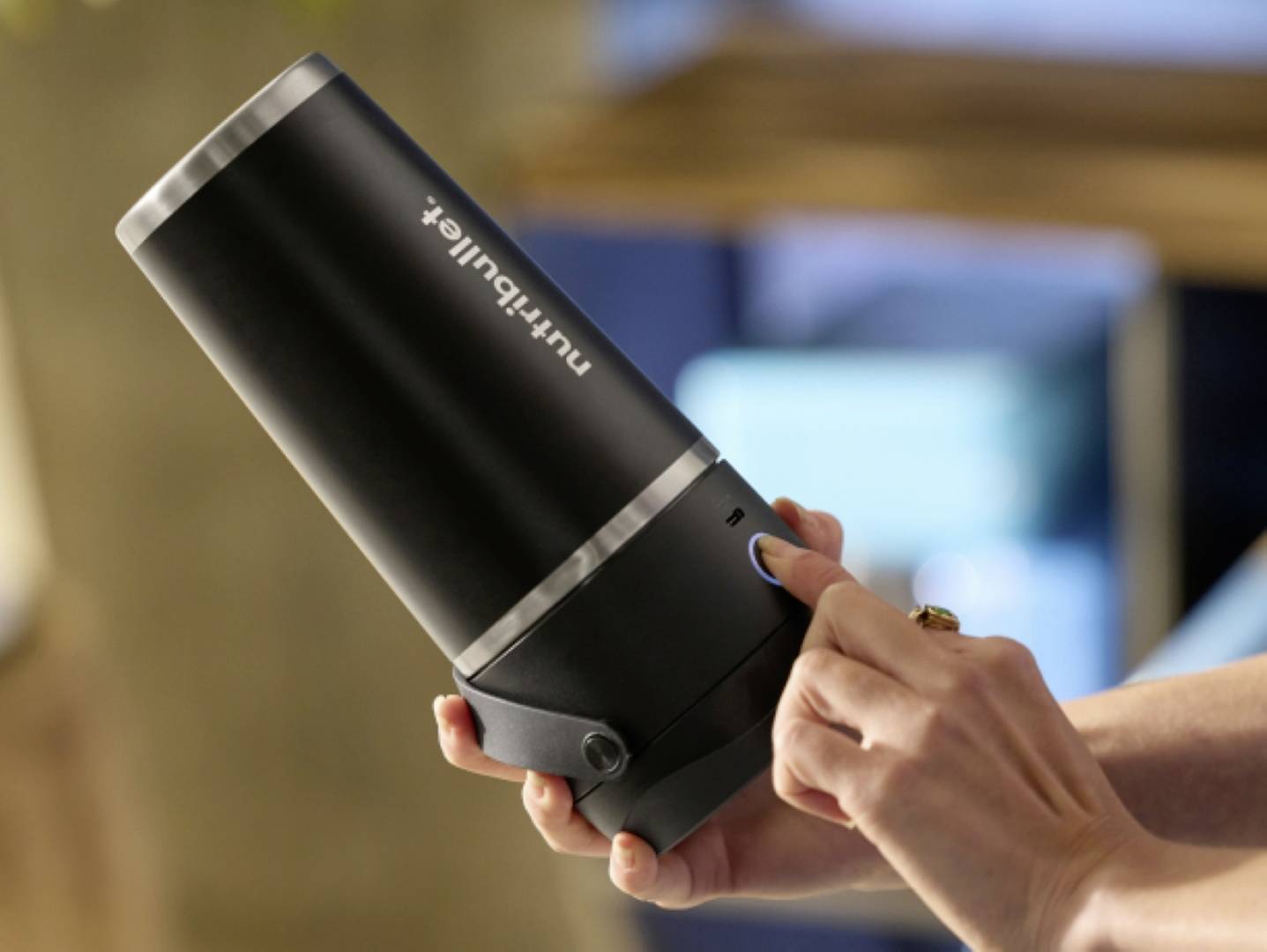 Osoba držiaca elegantný čierny prenosný mixér, ktorá stláča tlačidlo. Zariadenie je označené nápisom 'nutribullet'. Pozadie je jemne rozostreté.