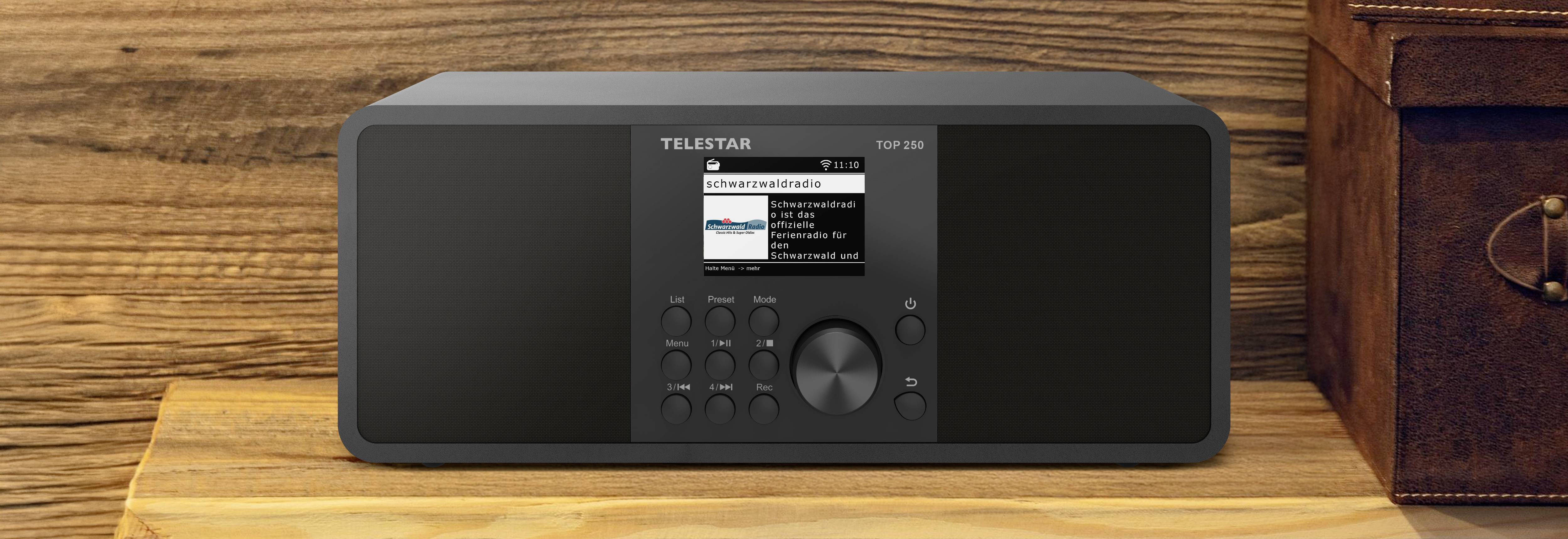 Na drevenej polici stojí čierne rádio TELSTAR TSP 90, ktoré má digitálny displej a ovládacie tlačidlá. Na displeji je zobrazená hudobná stopa.