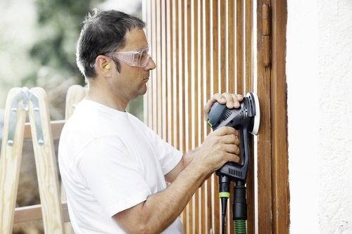 Osoba používa elektrickú brúsku na vyhladenie dreveného povrchu, má nasadené ochranné okuliare a chrániče sluchu, v pozadí je rebrík.