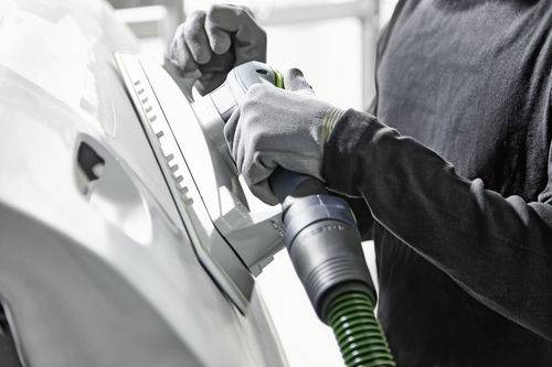 Osoba používajúca brúsku na autové dvere, oblečená v ochranných rukaviciach pre bezpečnosť. Dôraz je kladený na proces brúsenia, ktorý poukazuje na automechanickú opravu.