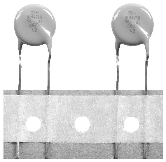 Dva diskové elektronické komponenty s dlhými vývodmi, označené '10N471K 5V', sú namontované na páske s perforovanými otvormi.
