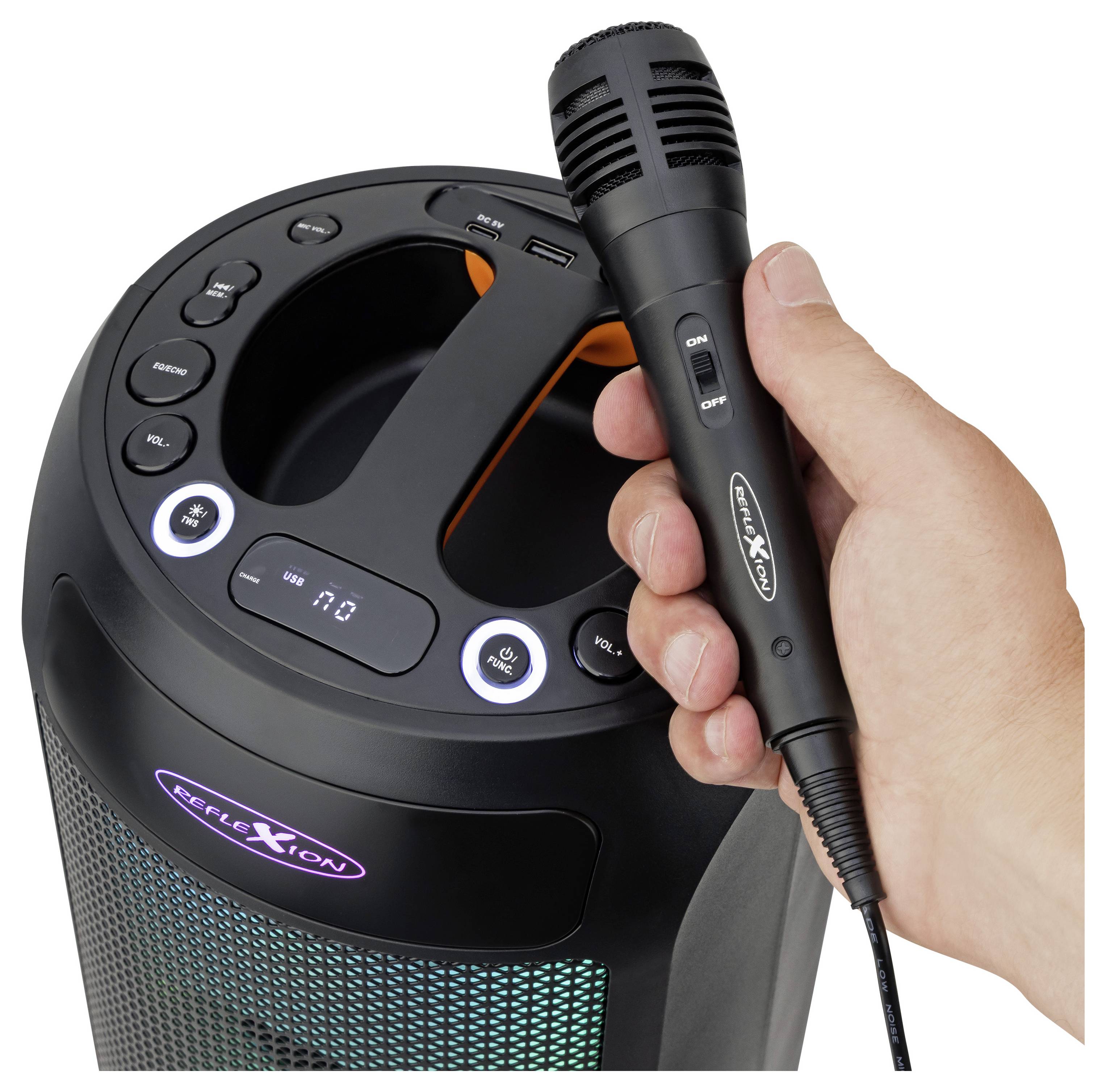 Ruka drží mikrofón pred reproduktorom s ovládacími tlačidlami a LED displejom, čo naznačuje zostavu pre karaoke.