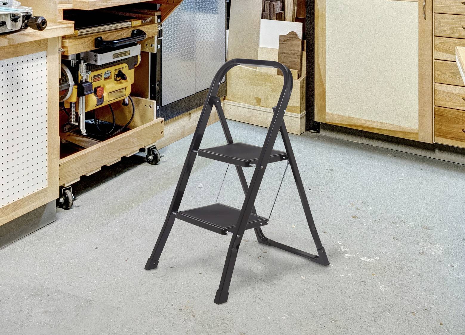Malý, čierny, dvojstupňový rebrík stojí na podlahe dielne obklopený drevenými skrinkami, pracovným stolom a rôznymi nástrojmi.