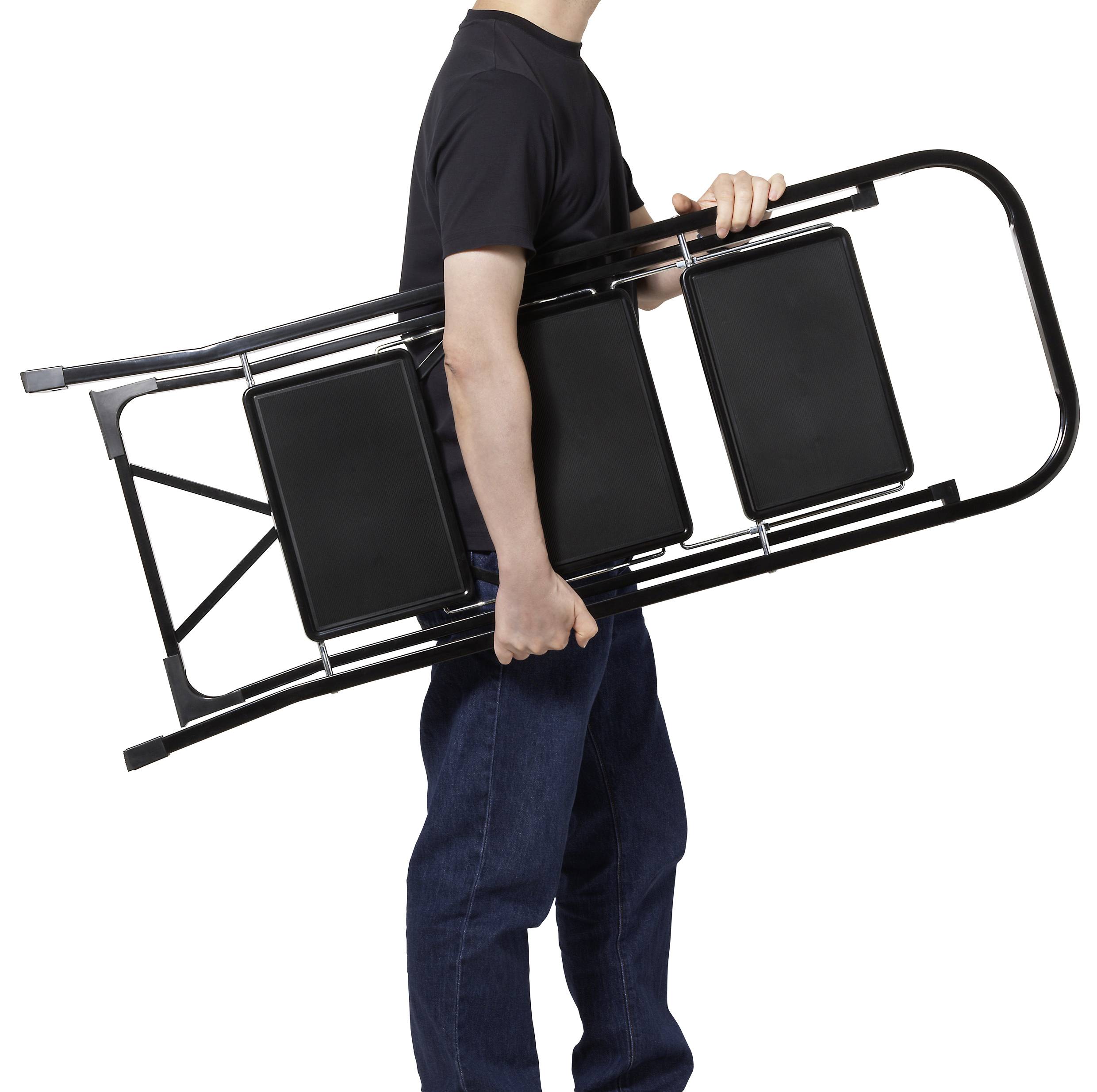 Osoba nesúca zloženú čiernu stoličku, oblečená v čiernej košeli a modrých džínsach, pred bielym pozadím.