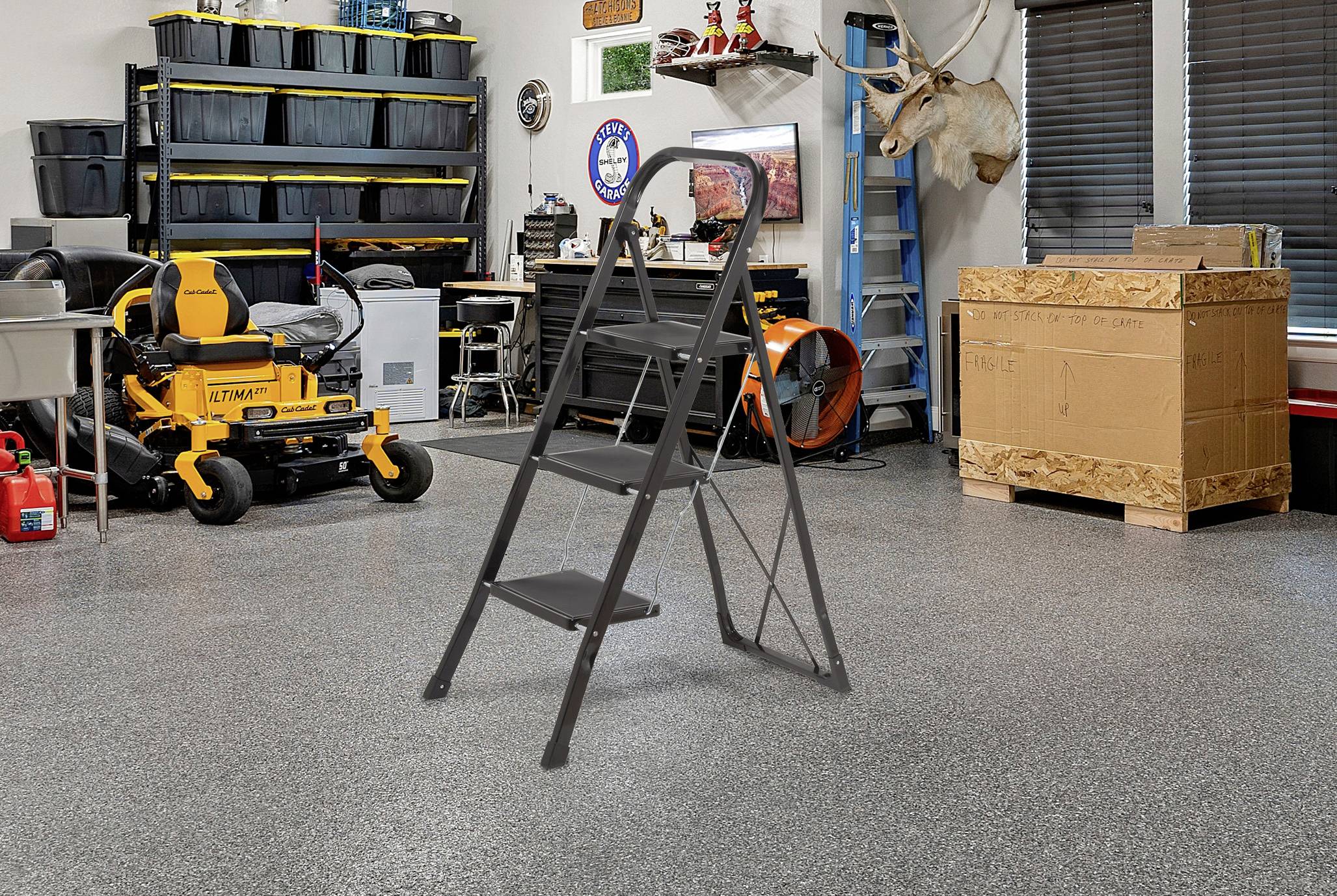 Sivý rebríkový rebrík je otvorený uprostred dobre organizovanej garáže s úložnými policami, kosačkou, namontovanou hlavou srny a veľkou kartónovou škatuľou.
