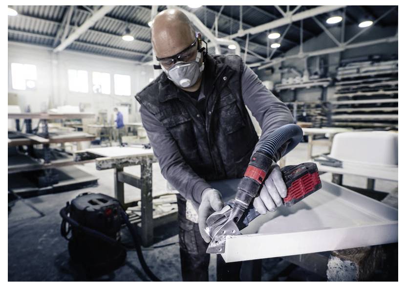 Osoba v bezpečnostnej výbave obsluhuje elektrický nástroj vo fabrike a tvaruje kus kovu. V pozadí sú viditeľné pracovné stoly a priemyselné zariadenia.