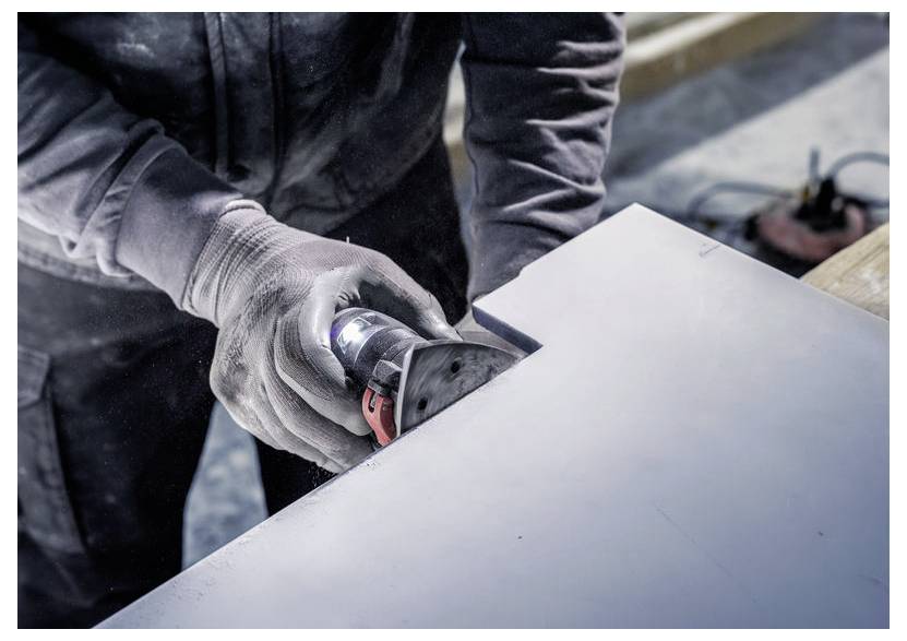 Osoba používa ručnú elektrickú brúsku na kus dreva, hladí jeho hrany v dielenskej atmosfére.