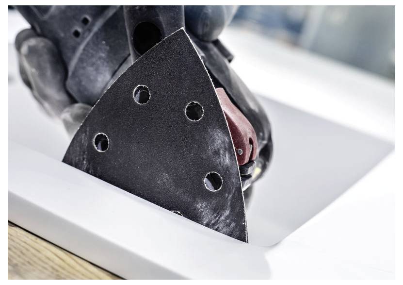 Ruka drží brúsku s trojuholníkovou brúsnou podložkou, ktorá vyhladzkuje hranu bieleho povrchu, pravdepodobne kusu nábytku alebo pracovnej dosky.