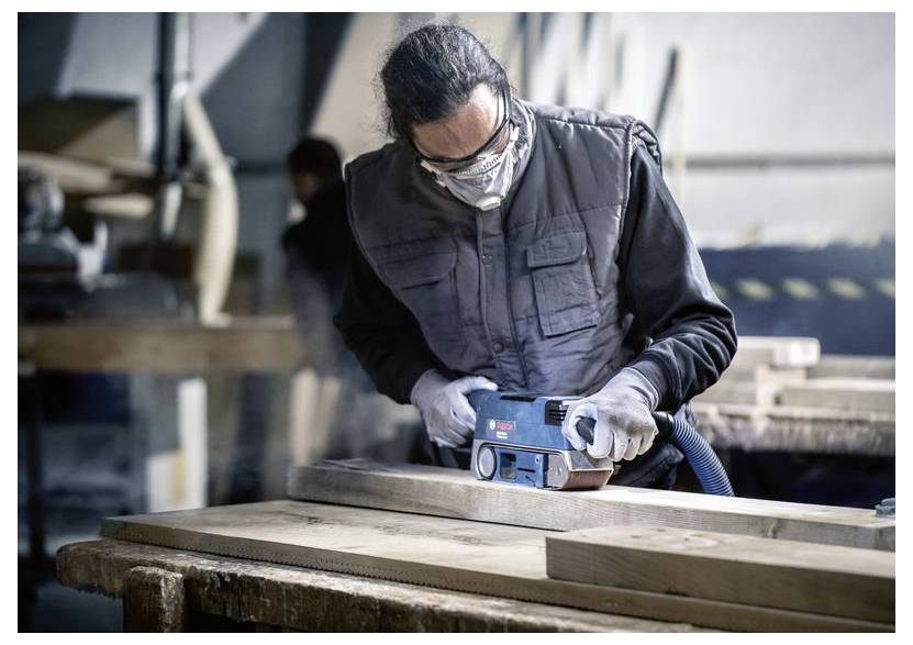 Osoba v bezpečnostnej výbave obsluhuje elektrickú brúsku na drevenom doske v dielni a sústreďuje sa na svoju prácu.