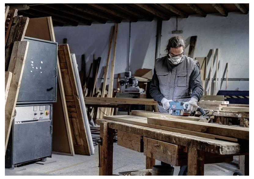Osoba v ochranných pomôckach obsluhuje brúsku na drevené dosky v stolárskej dielni s drevom a vybavením v pozadí.