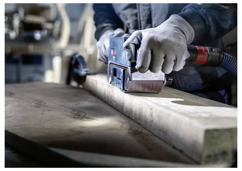 Osoba v rukaviciach používa elektrickú brúsku na vyhladenie drevenej dosky v dielni so zameraním na presnosť a remeselnú zručnosť.