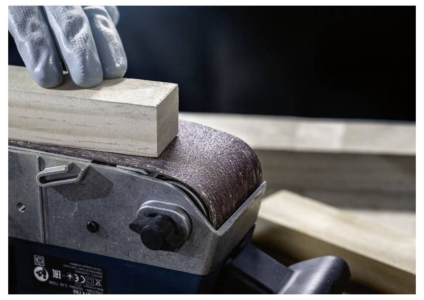 Rukavicou oblečená ruka drží drevený blok pritlačený na pásovú brúsku, ktorá vyhladkuje jeho povrch. Je viditeľná pilina, čo naznačuje prebiehajúcu brúsnu prácu.