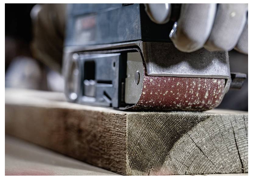 Detailný záber na elektrickú brúsku, ktorá vyhladkuje povrch drevenej dosky. Brúsny pás je v pohybe a odstraňuje piliny z dreva.