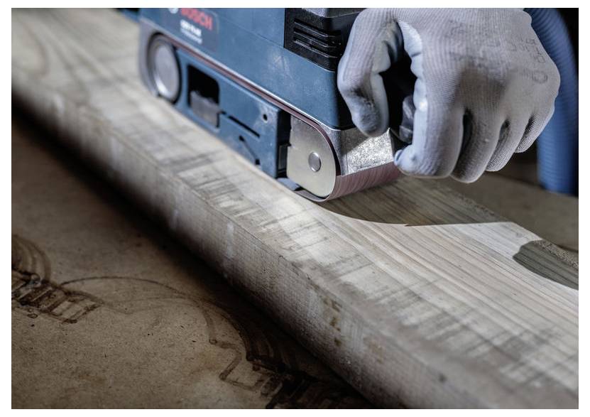 Osoba používa elektrickú pásovú brúsku na drevenej ploche, vyhladkávajúc drevo. Ruka je chránená ochrannou rukavicou.