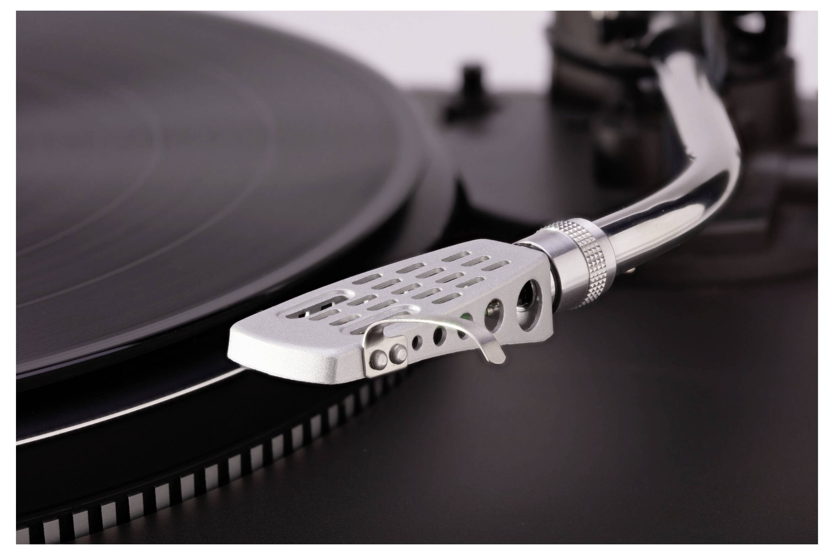 Detailný záber ramena gramofónu a stylusa umiestnených nad otáčajúcim sa čiernym vinylom, zvýrazňujúci drážky a kovové detaily.