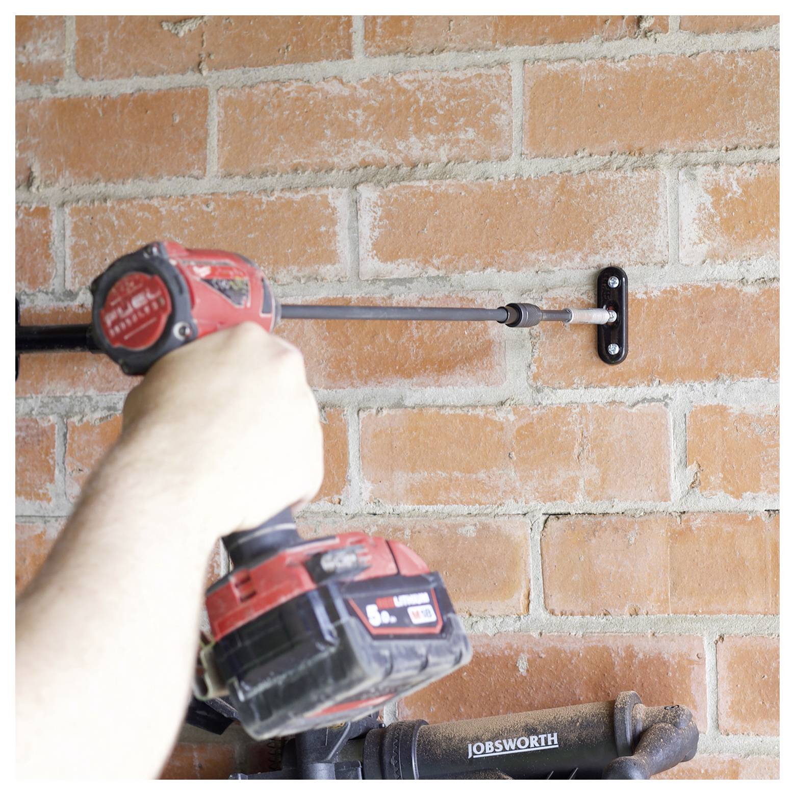 Osoba používajúca červenú elektrickú vŕtačku na pripevnenie čierneho stenového držiaka k tehlovej stene.