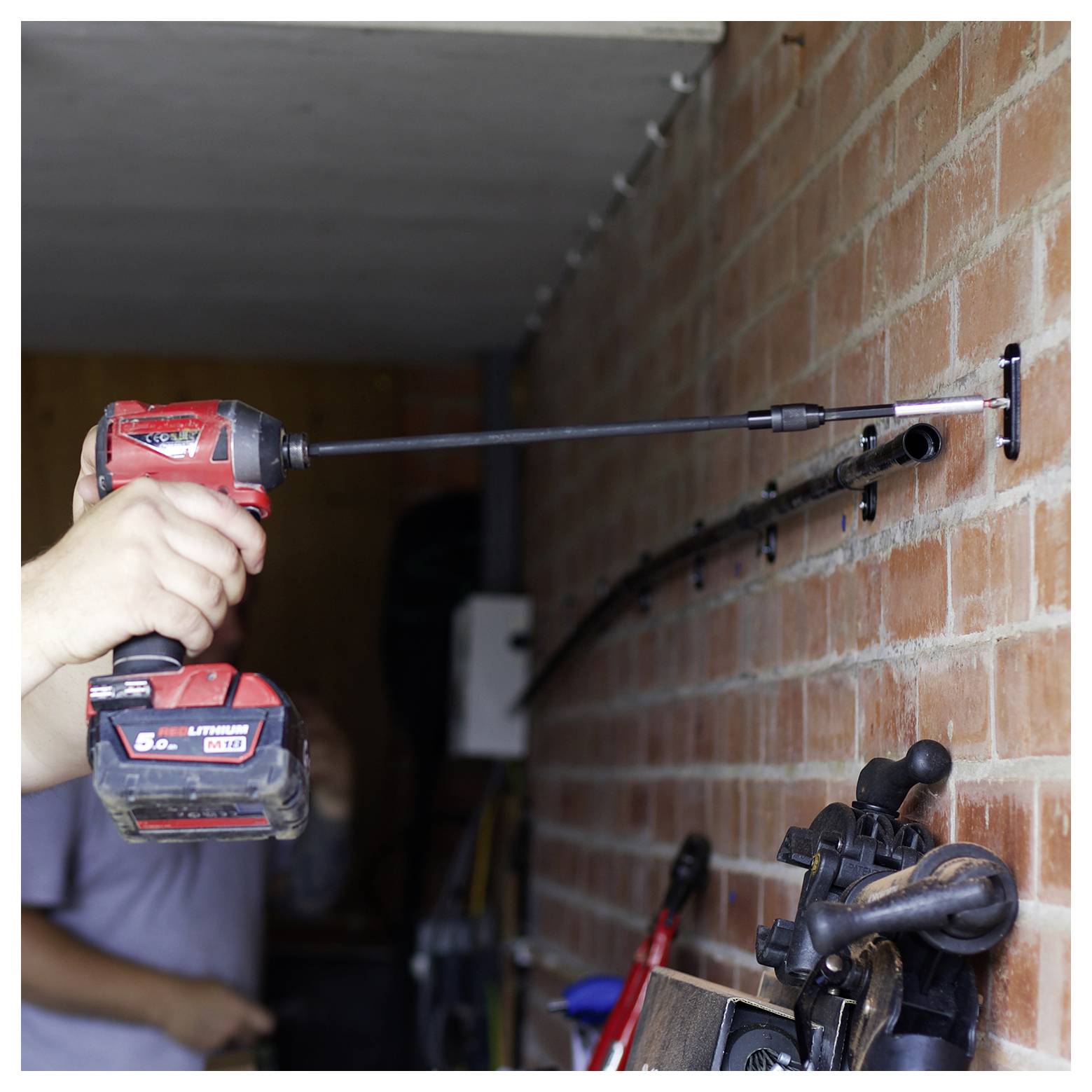 Osoba používajúca červenú bezdrôtovú vŕtačku na upevnenie kovovej tyče na tehlovú stenu, uprostred rôznych ručných nástrojov uložených na polici.