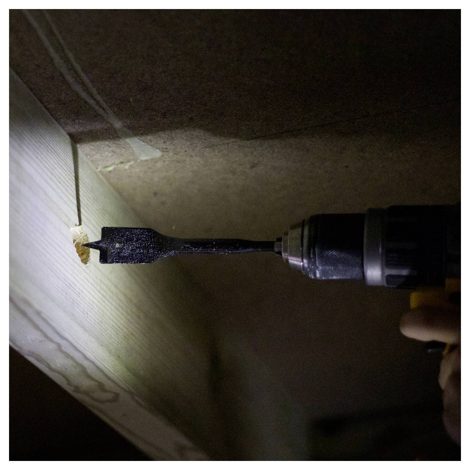 Osoba používa vŕtačku na vytvorenie diery do drevenej dosky, s vrtákom na drevo a tlmeným svetlom v pracovnej oblasti.