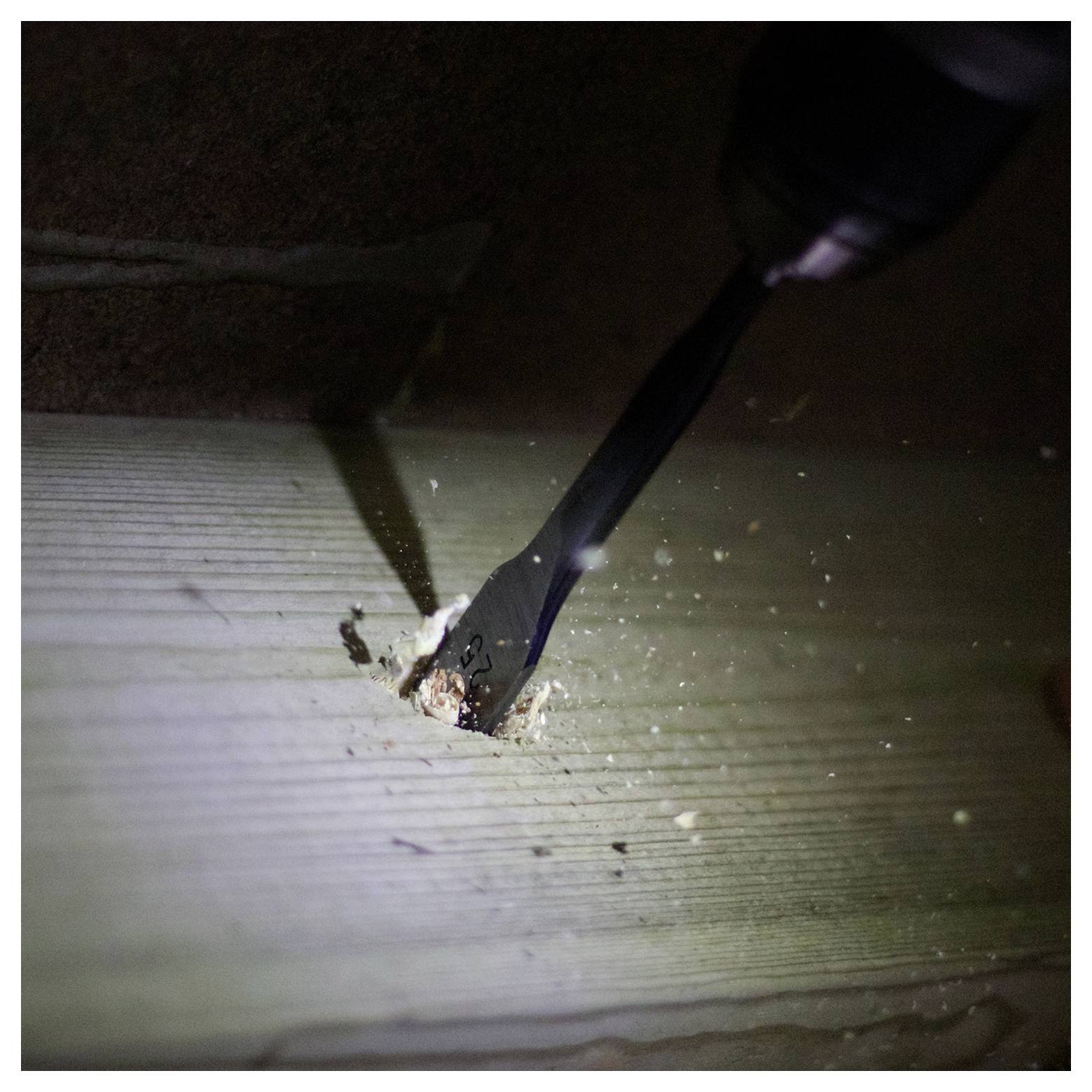 Vŕtačka vytvárajúca otvor cez drevenú plochu, s drevnými trieskami roztrúsenými okolo, čo naznačuje prebiehajúcu stavebnú alebo stolársku prácu.