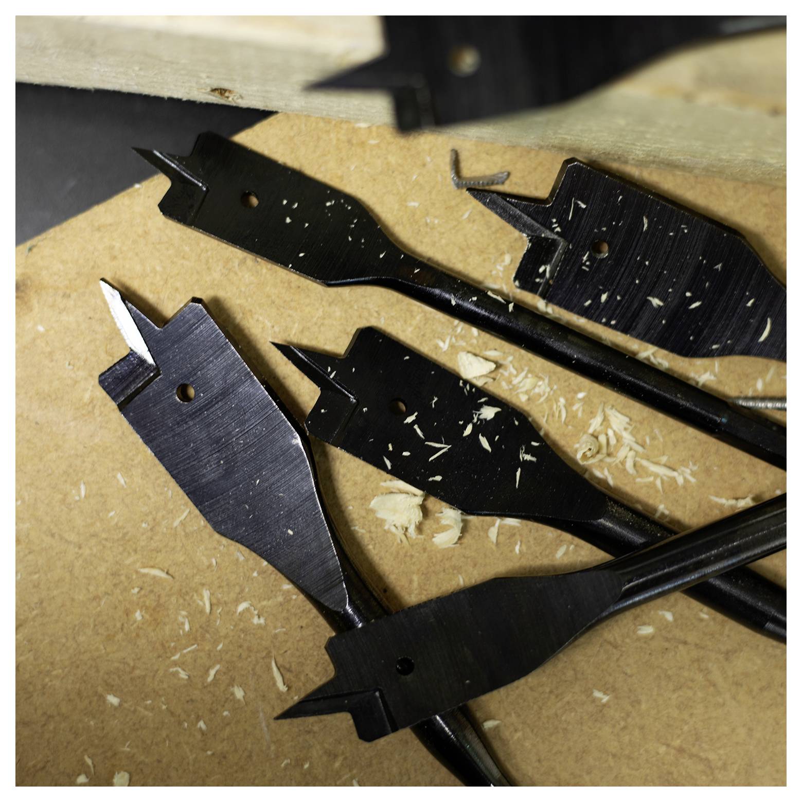 Ploché drevené vrtáky roztrúsené na drevenej ploche s trieskami dreva. Niektoré vrtáky vykazujú známky použitia, pričom jeden vrták je čiastočne na okraji.